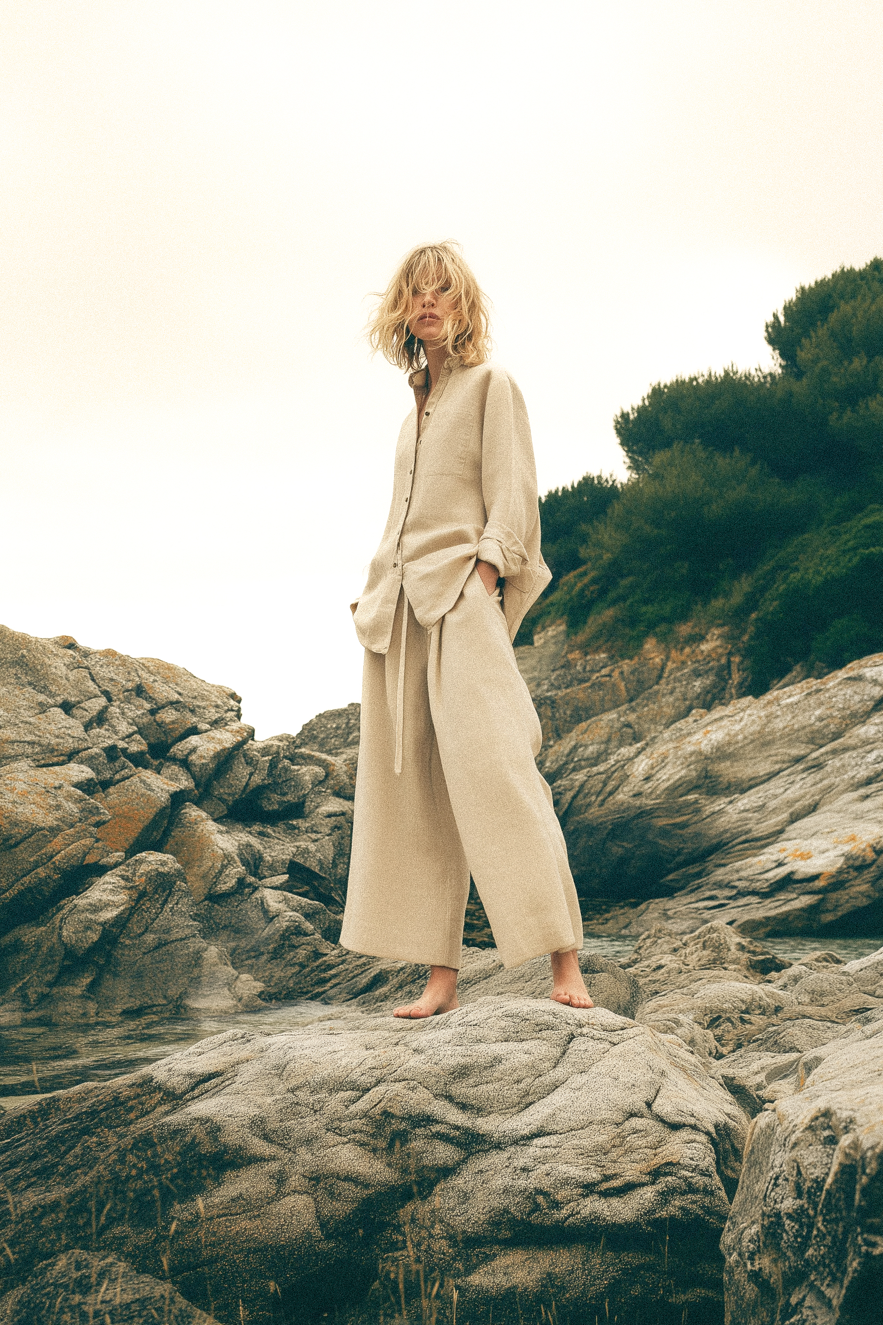 Adult woman in linen shirt and pants standing on rocks in a coastal environment on a cloudy day, fashion editorial.