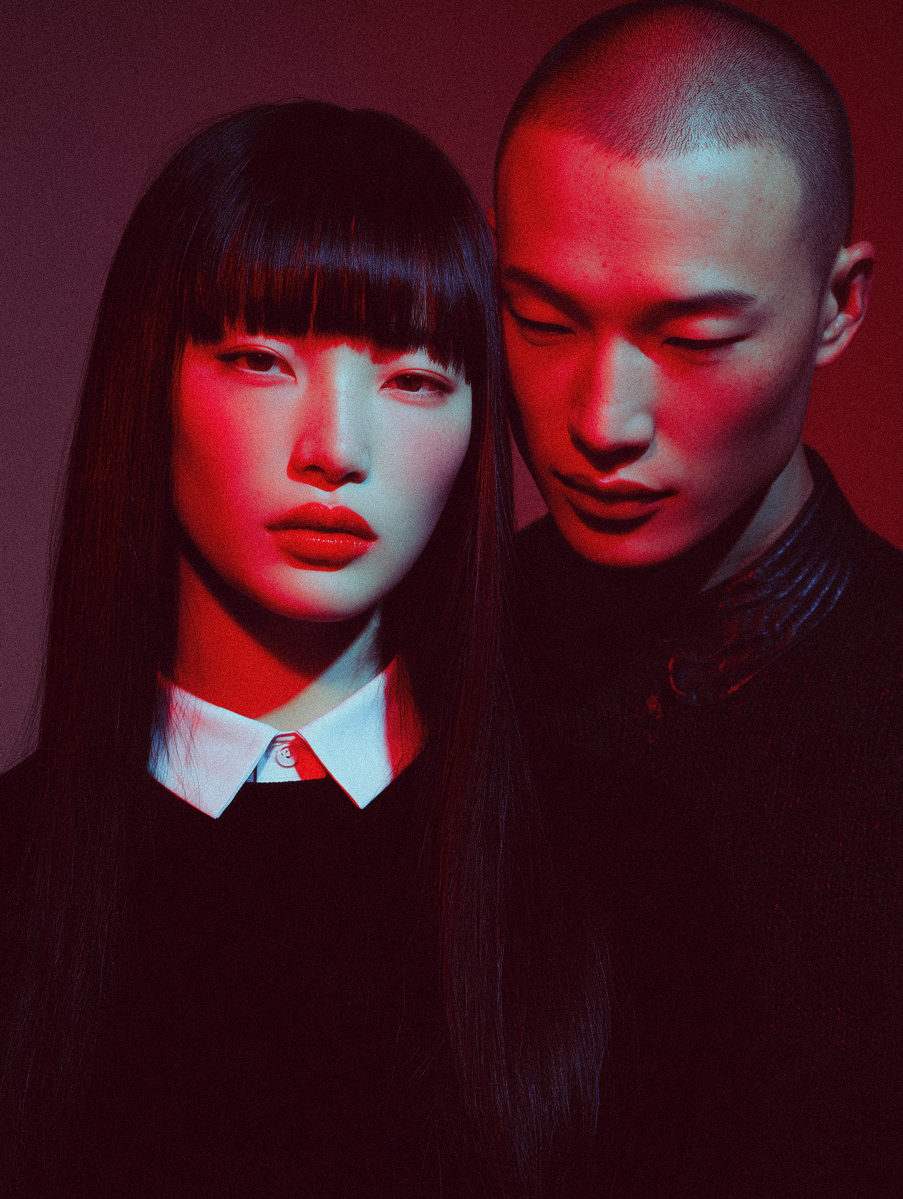 Portrait of an Asian female model with dark hair and red lipstick with a shaved head male model in a close-up shot against a red background.