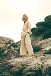 Barefoot woman in an oversized beige linen shirt and wide-leg trousers standing on coastal rocks.