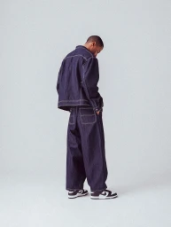 Man wearing a dark blue denim jacket and matching trousers with contrast white stitching against a light grey studio background.