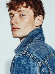 Close-up portrait of a young man with red hair and freckles wearing a blue denim jacket against a plain background.