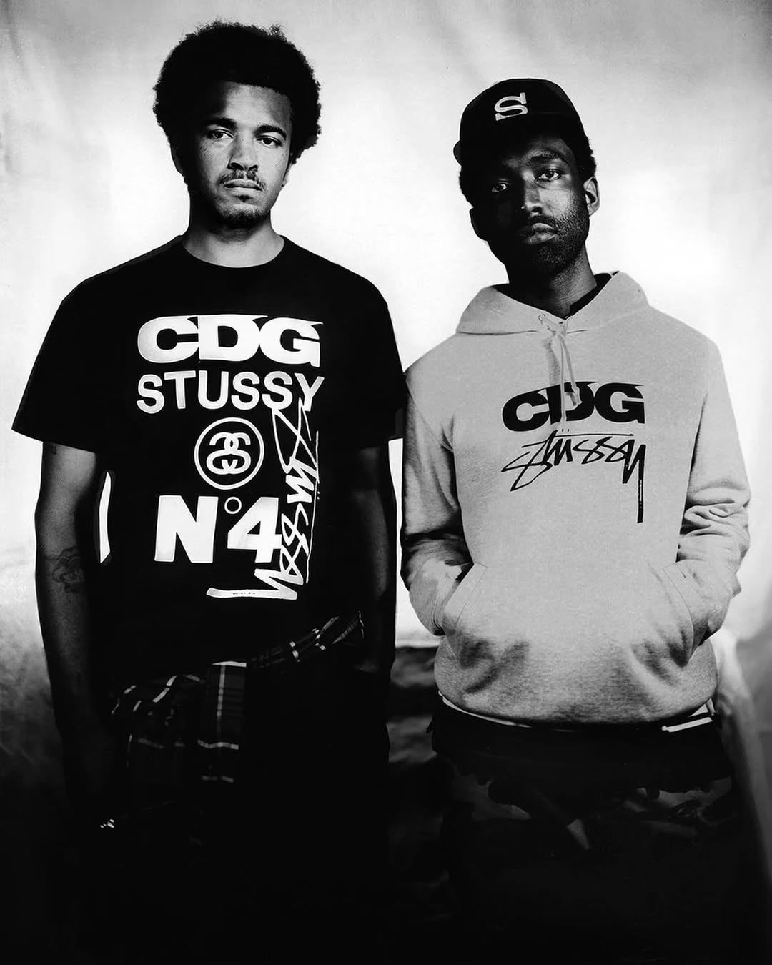 Two adult men pose indoors wearing branded streetwear t-shirts and hoodies in a black and white studio fashion shot.