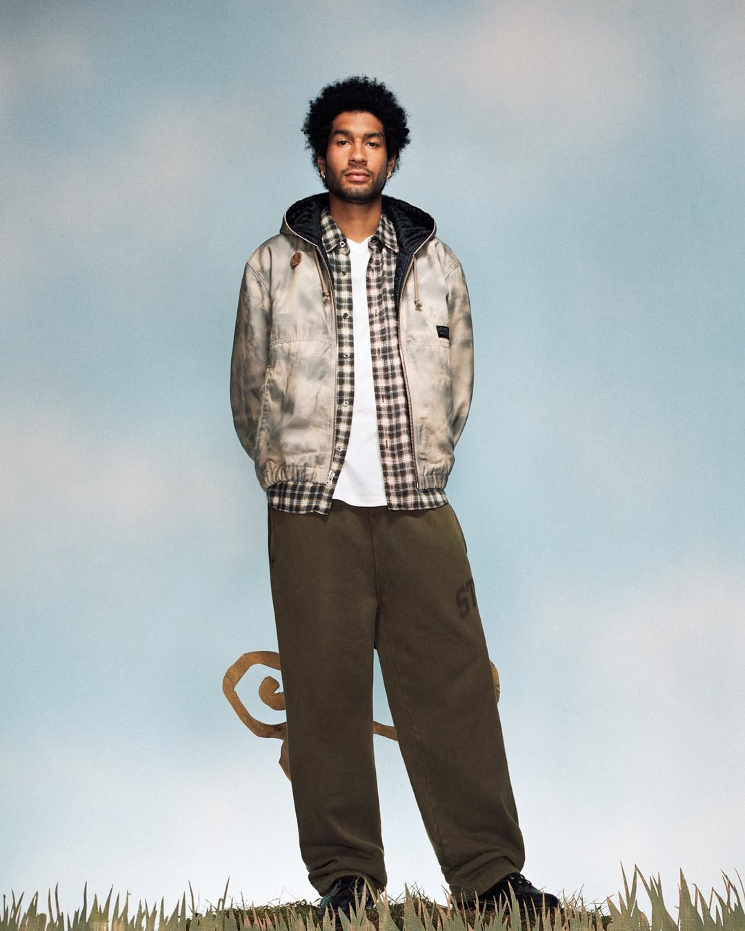 Young Black male model in a hooded jacket and olive pants poses against a light blue sky backdrop with whimsical golden accents.