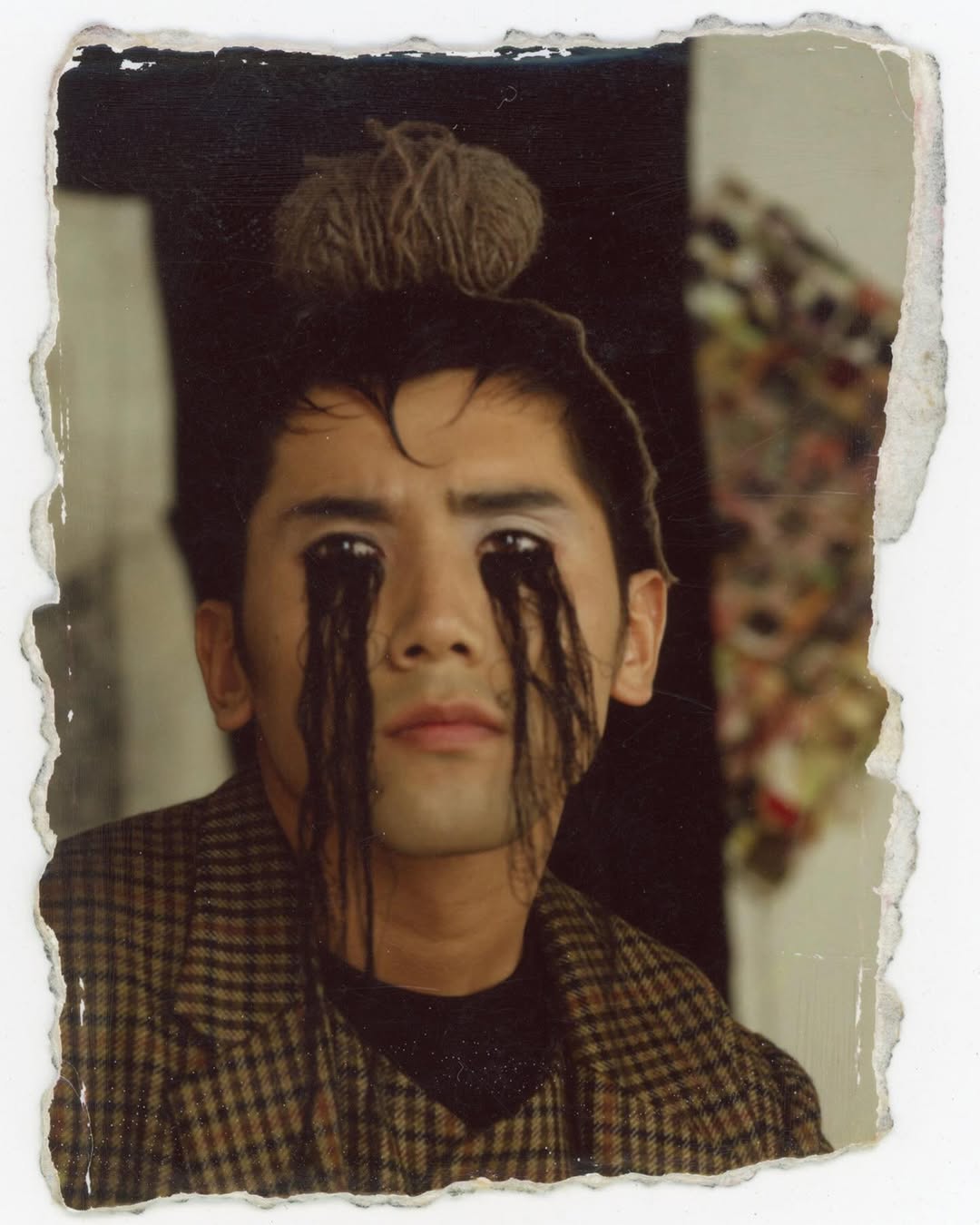 Artistic portrait of an adult with dark hair or yarn strands over their eyes, wearing a brown patterned blazer, and yarn ball on their head.