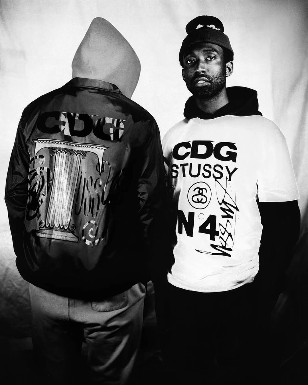Two people in black and white; one man faces forward wearing a beanie and T-shirt with graphic text. The other wears a jacket.