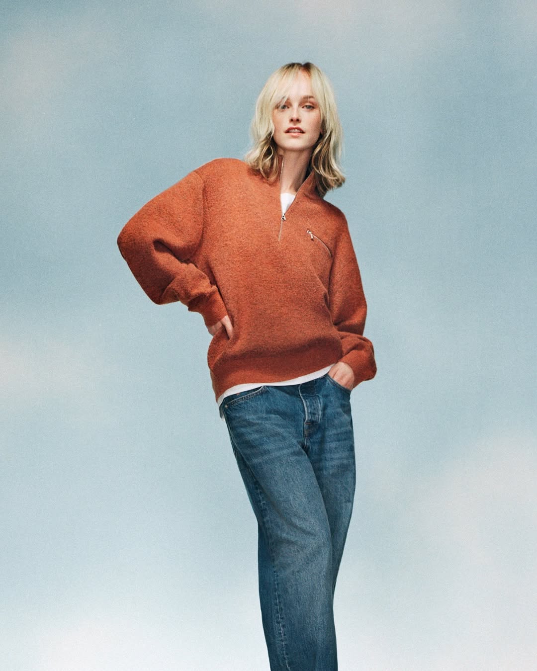 An adult female model poses in a studio with a light blue background wearing a rust-colored quarter-zip sweatshirt and jeans, looking at the camera.