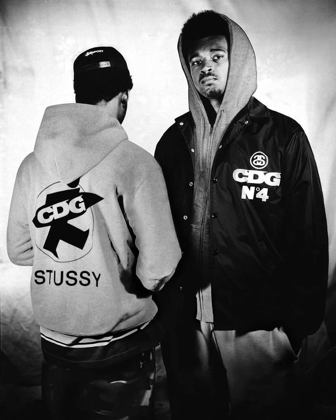 Two stylish adult males pose indoors wearing streetwear hoodies and jackets featuring logos in a black and white fashion photograph.