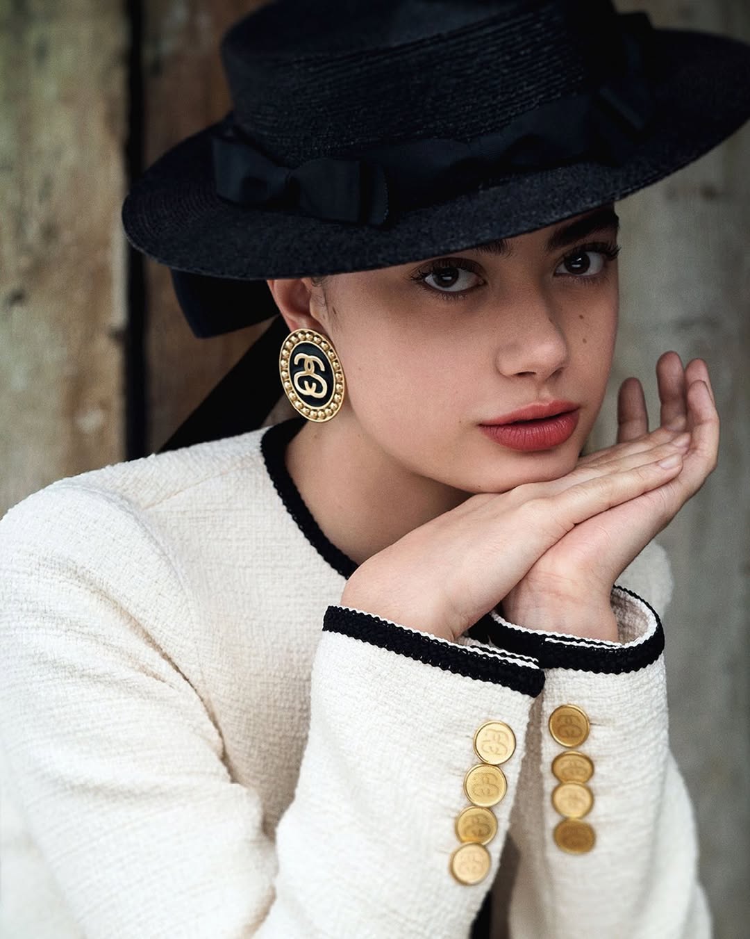 A young adult woman in a cream-colored jacket and black hat with a ribbon posing in a close-up shot against a neutral backdrop.