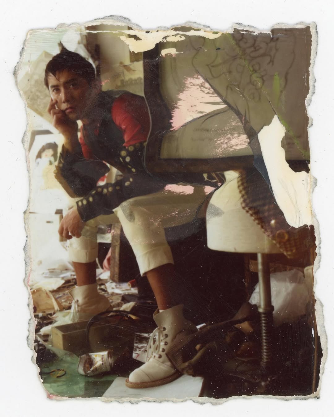 An eye-level indoor portrait shows an adult man seated with a hand to his face, wearing white pants, boots, and a black jacket with red shoulders.
