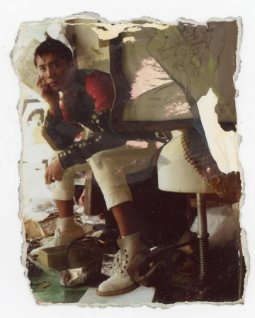 An eye-level indoor portrait shows an adult man seated with a hand to his face, wearing white pants, boots, and a black jacket with red shoulders.