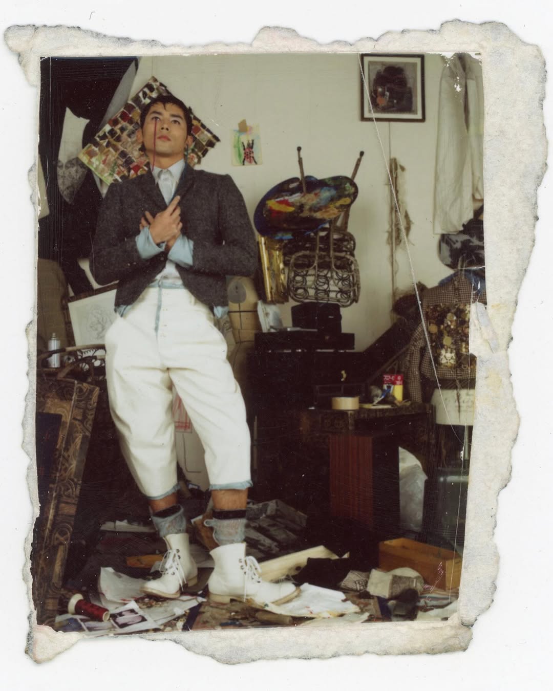 Full body shot of an androgynous person with dark hair wearing a grey blazer, light blue shirt, and white pants posing indoors in a messy room.