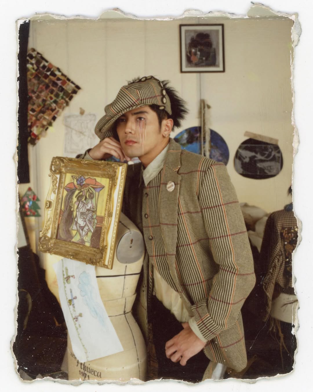 A stylish person wearing a plaid suit and cap leans on a mannequin with an artistic portrait, in a room with paintings and designs.