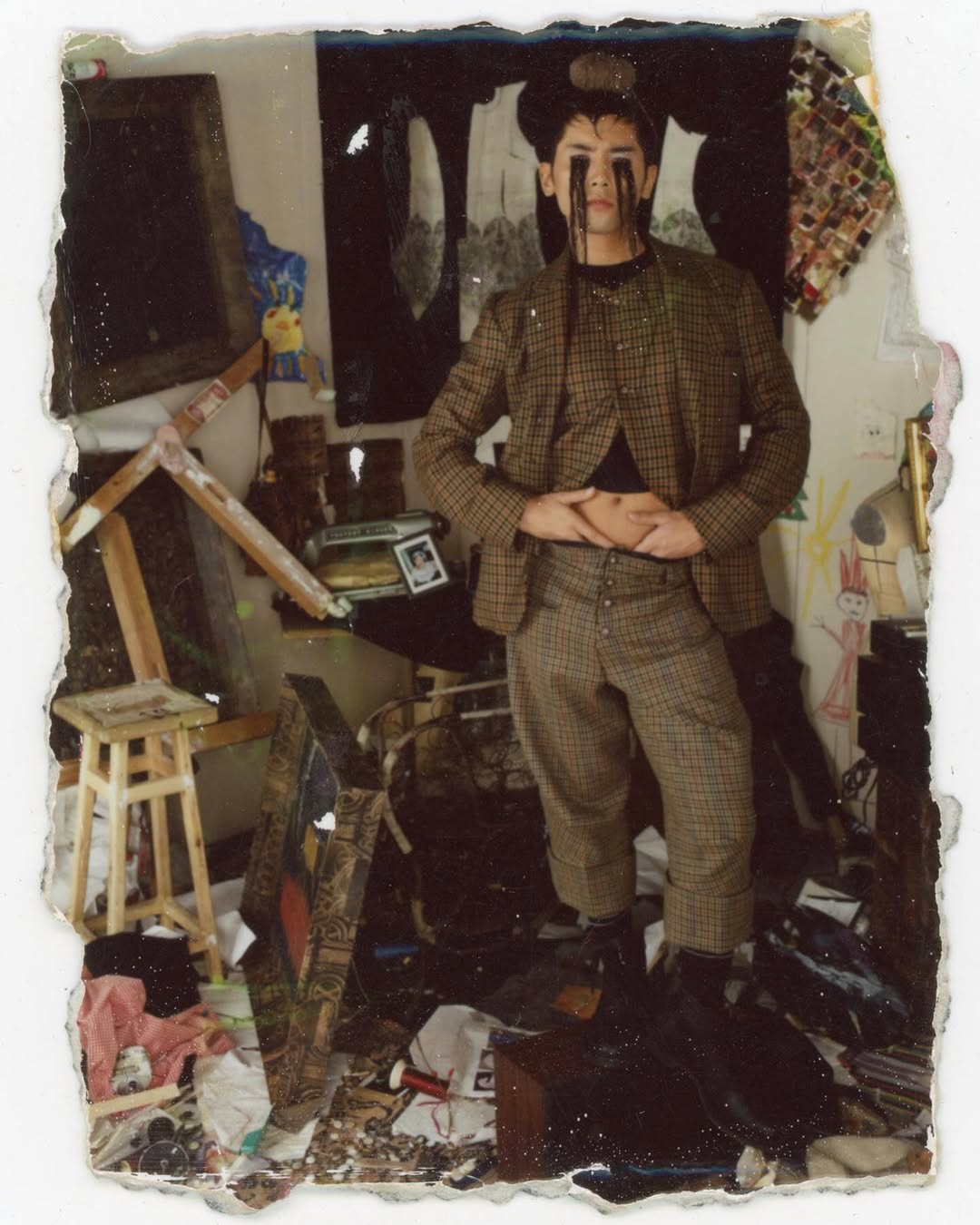Person in a patterned suit with dark makeup stands in a cluttered interior space with art supplies and various objects scattered around.