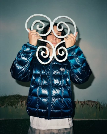 Person in puffer jacket holds up decorative, white, spiral design. Indoor portrait with soft lighting. Neutral background.