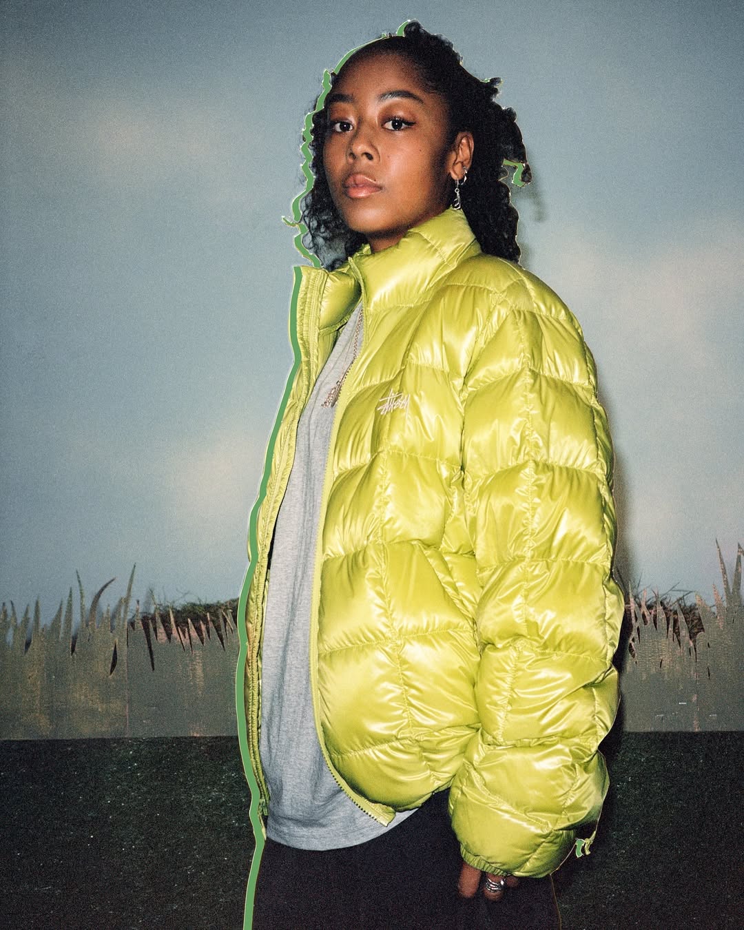 An adult female with curly hair poses in a shiny yellow puffer jacket against a painted backdrop, looking directly at the camera.