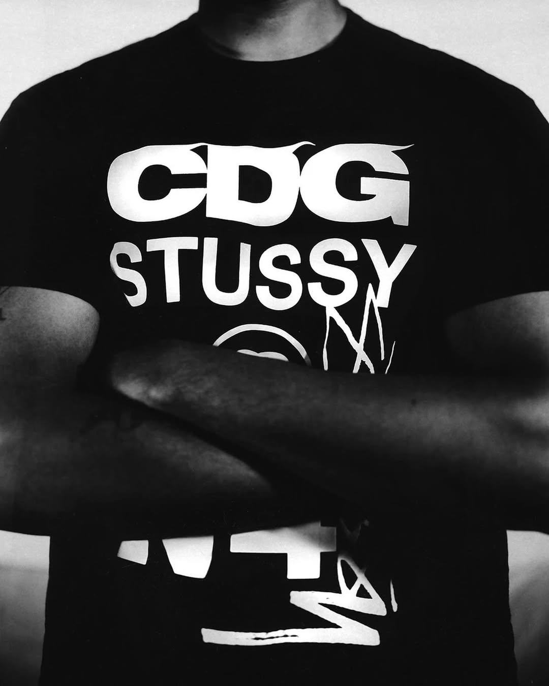 Black and white portrait of a person wearing a black t-shirt with white 'CDG Stussy' text and arms crossed in front of a white background.