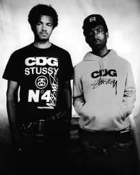 Black and white portrait of two Black men wearing graphic t-shirts and hoodies against a textured studio background.