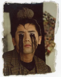 Portrait of a man wearing a plaid blazer with dark strands streaming from his eyes, photographed by Mark Lebon.