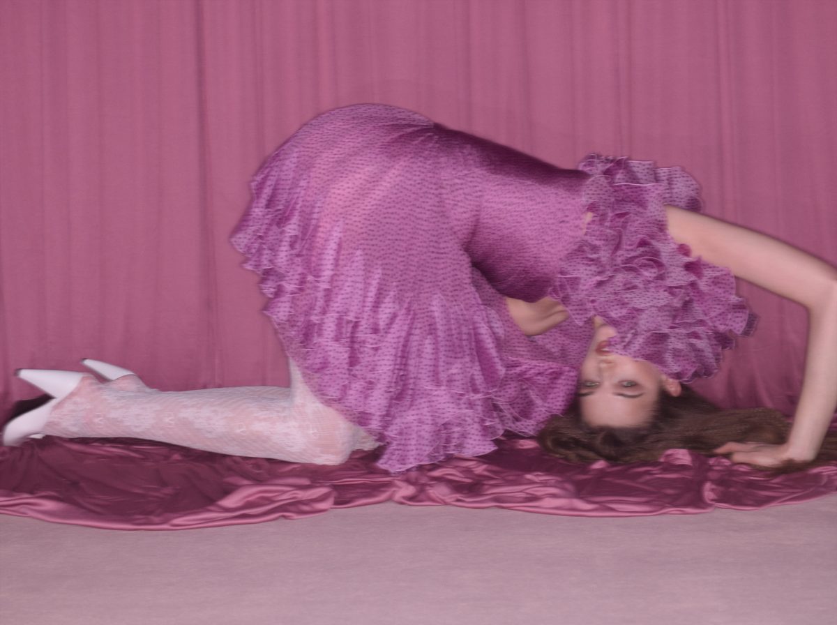 A woman in a purple dress and white stockings poses on a plum-colored fabric in front of a matching purple curtain backdrop.