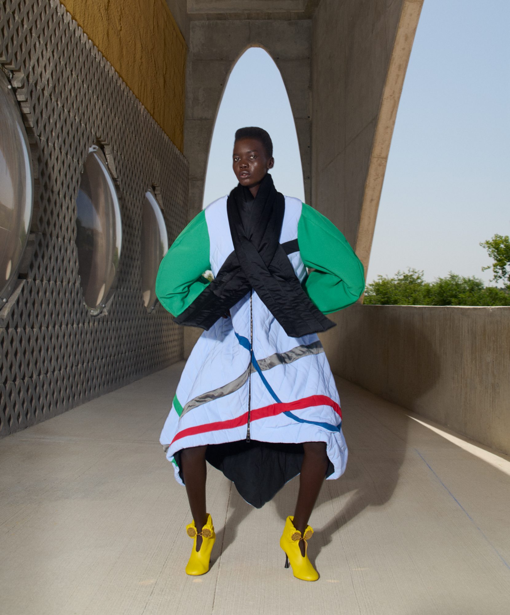 A statuesque figure in a color-blocked coat and bright yellow shoes stands confidently in a minimalist architectural space.