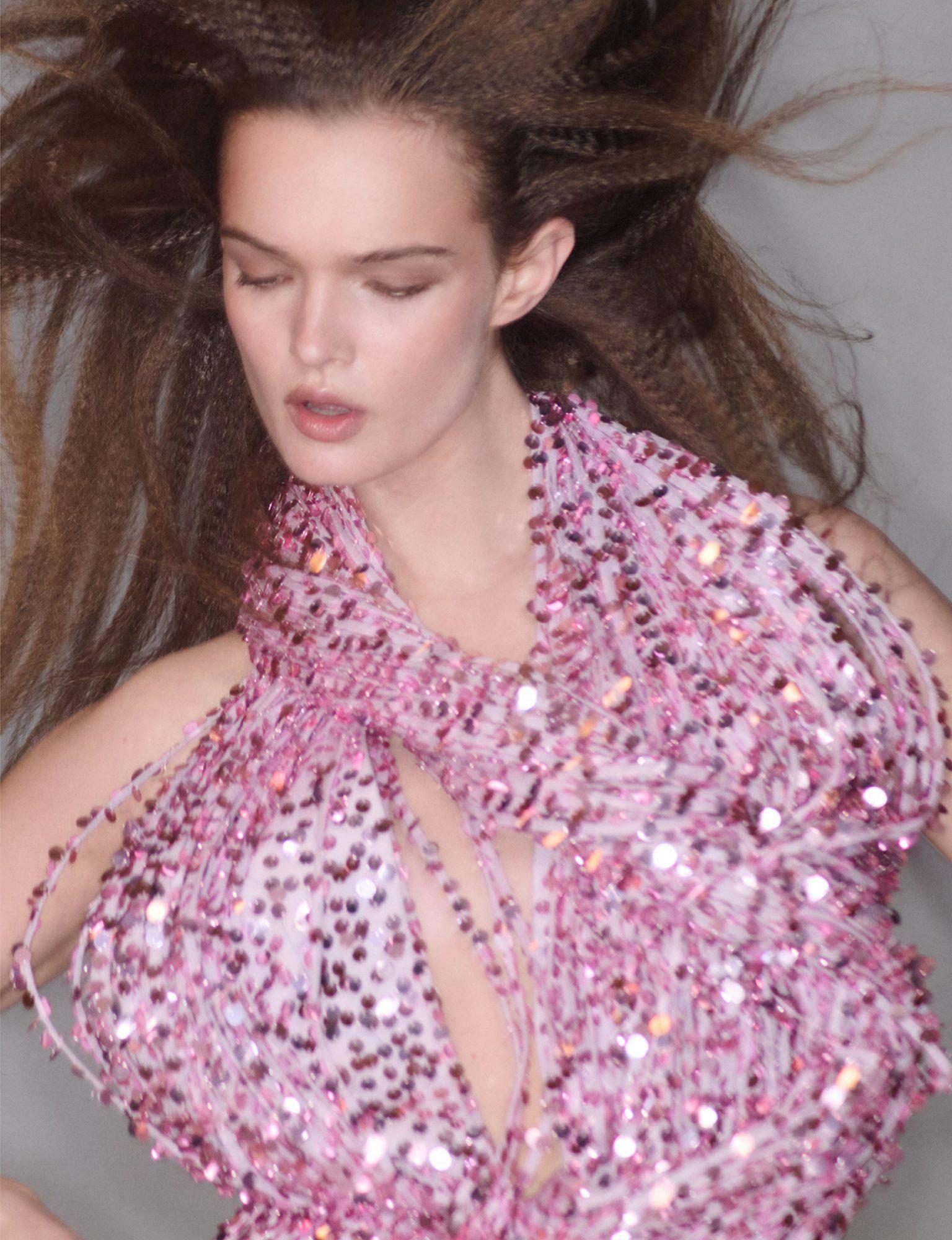 Close-up of a female model with crimped long brown hair wearing a sparkling pink beaded halter top, posing in a photo studio.