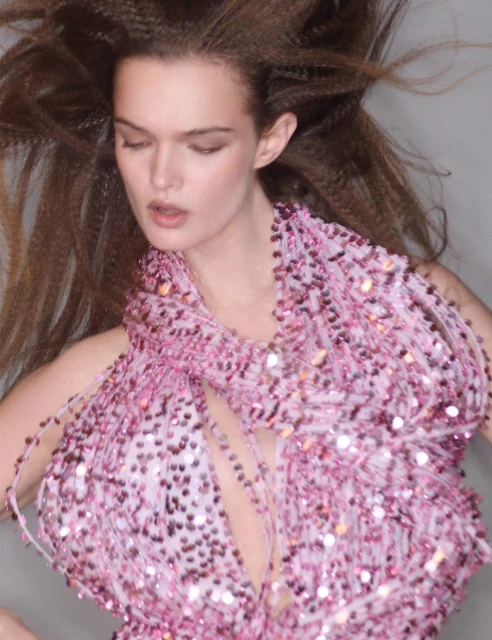 Close-up of a female model with crimped long brown hair wearing a sparkling pink beaded halter top, posing in a photo studio.