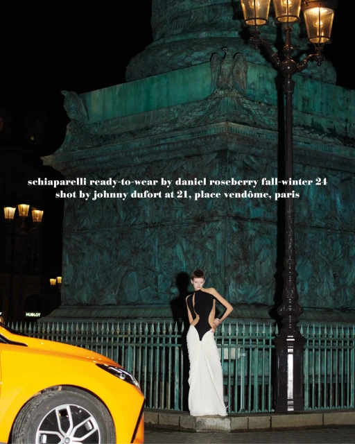 Fashion shot of a woman in an evening gown posing in front of a monument at night, with a blurred yellow taxi in the foreground.
