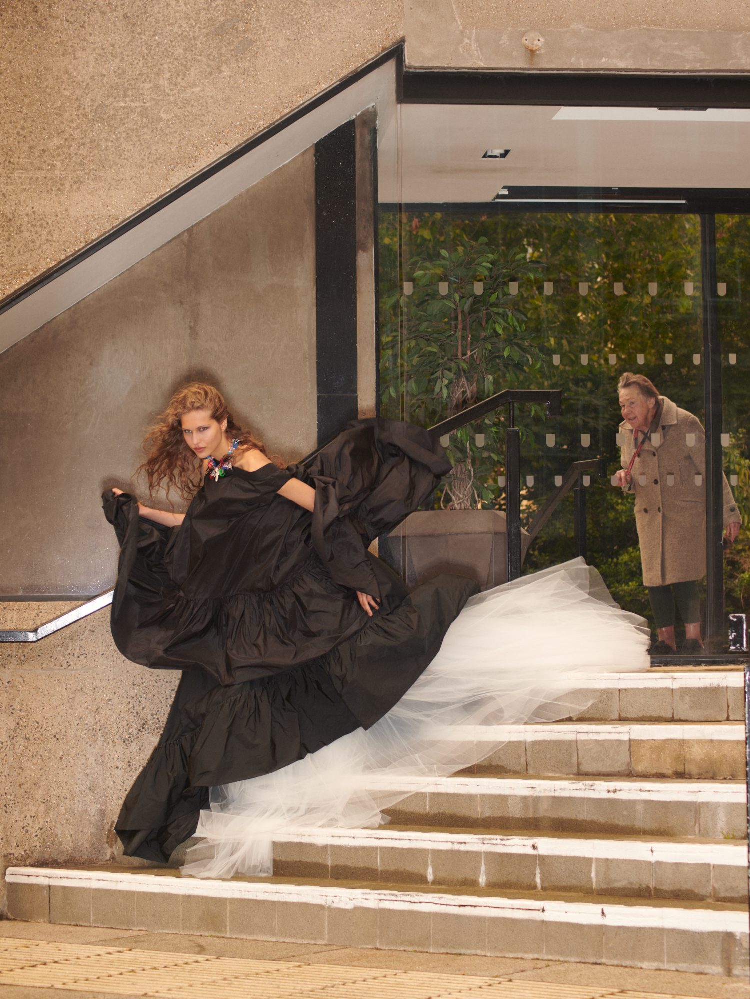 Fashion shot of a woman on stairs in a black gown with a white tulle train as an older woman looks on through a glass doorway.