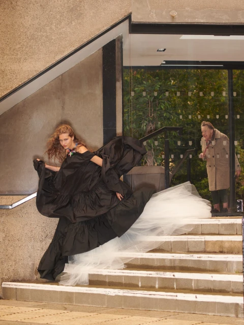 Fashion shot of a woman on stairs in a black gown with a white tulle train as an older woman looks on through a glass doorway.