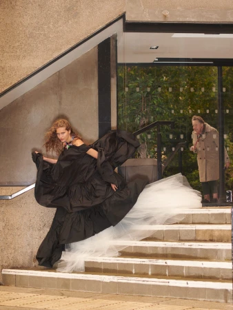 Fashion shot of a woman on stairs in a black gown with a white tulle train as an older woman looks on through a glass doorway.