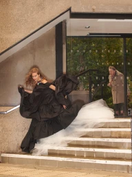 Model in voluminous black and white gown running down outdoor concrete stairs past an elderly person standing in a glass doorway.