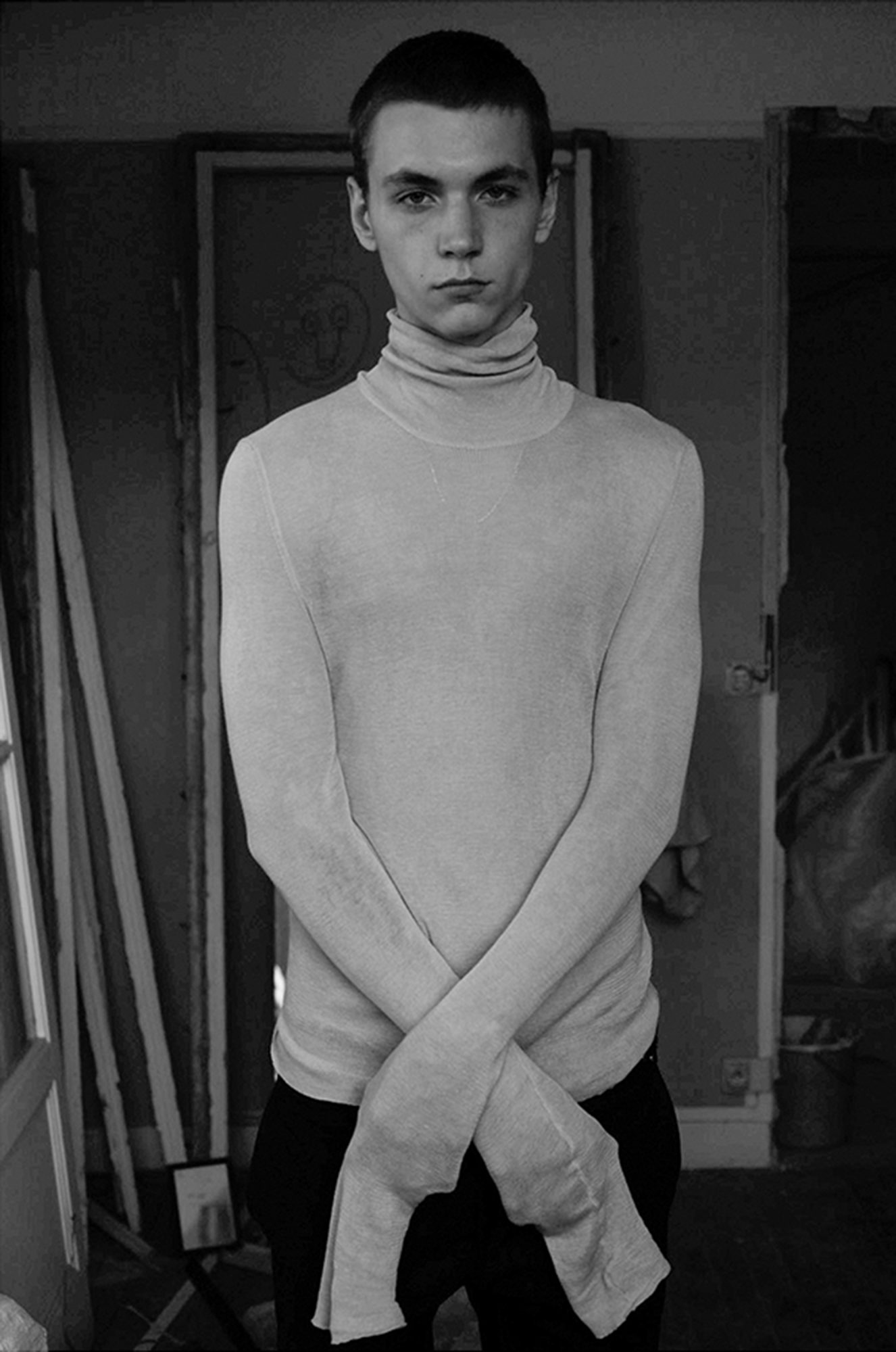 Monochrome portrait of a young adult male with short hair, wearing a turtleneck sweater, arms crossed, against a blurred background.