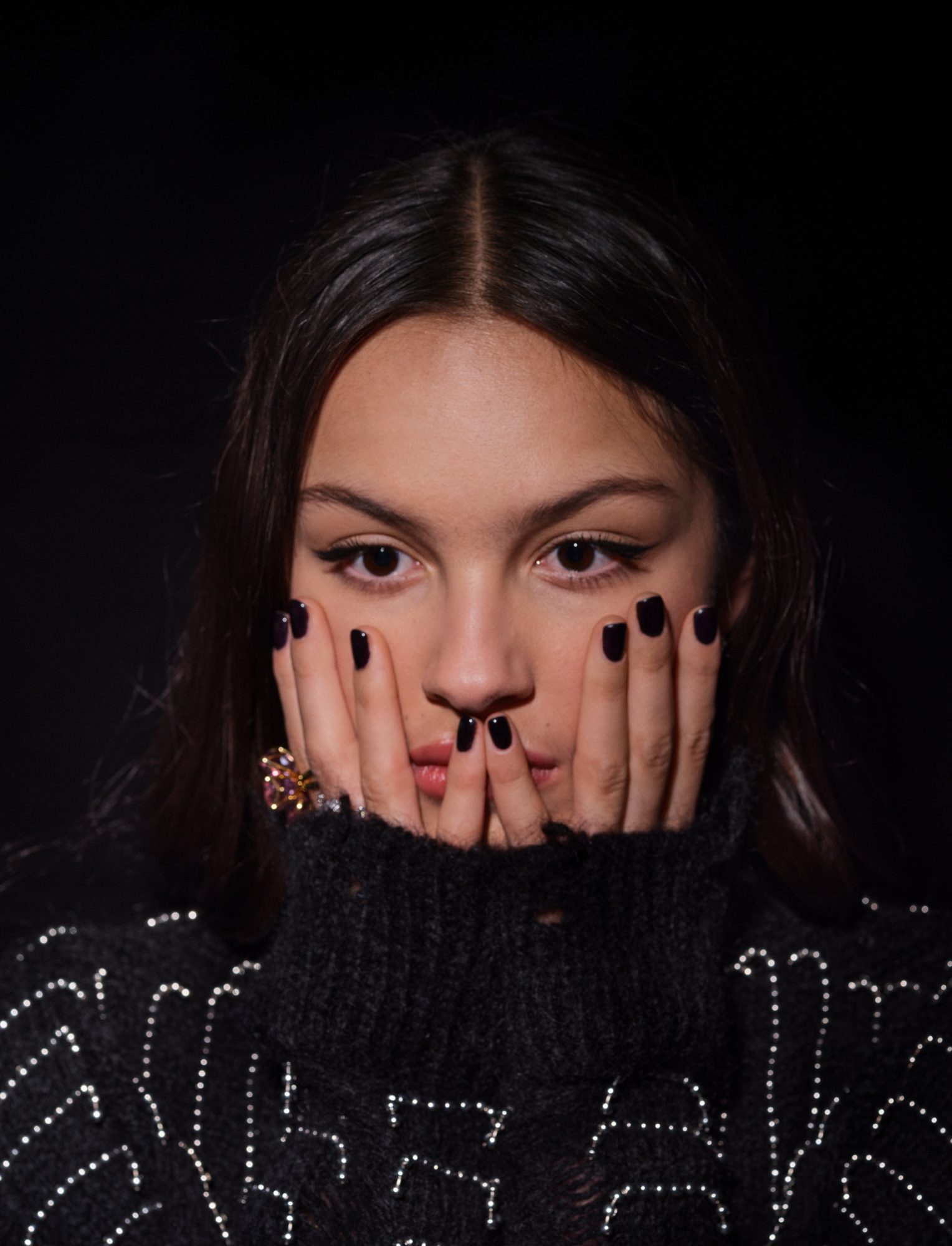 Close-up portrait of a woman with dark hair and hands near her face wearing a black turtleneck sweater in front of a black background.