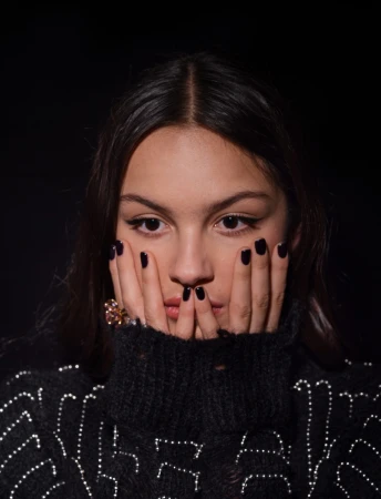 Close-up portrait of a woman with dark hair and hands near her face wearing a black turtleneck sweater in front of a black background.