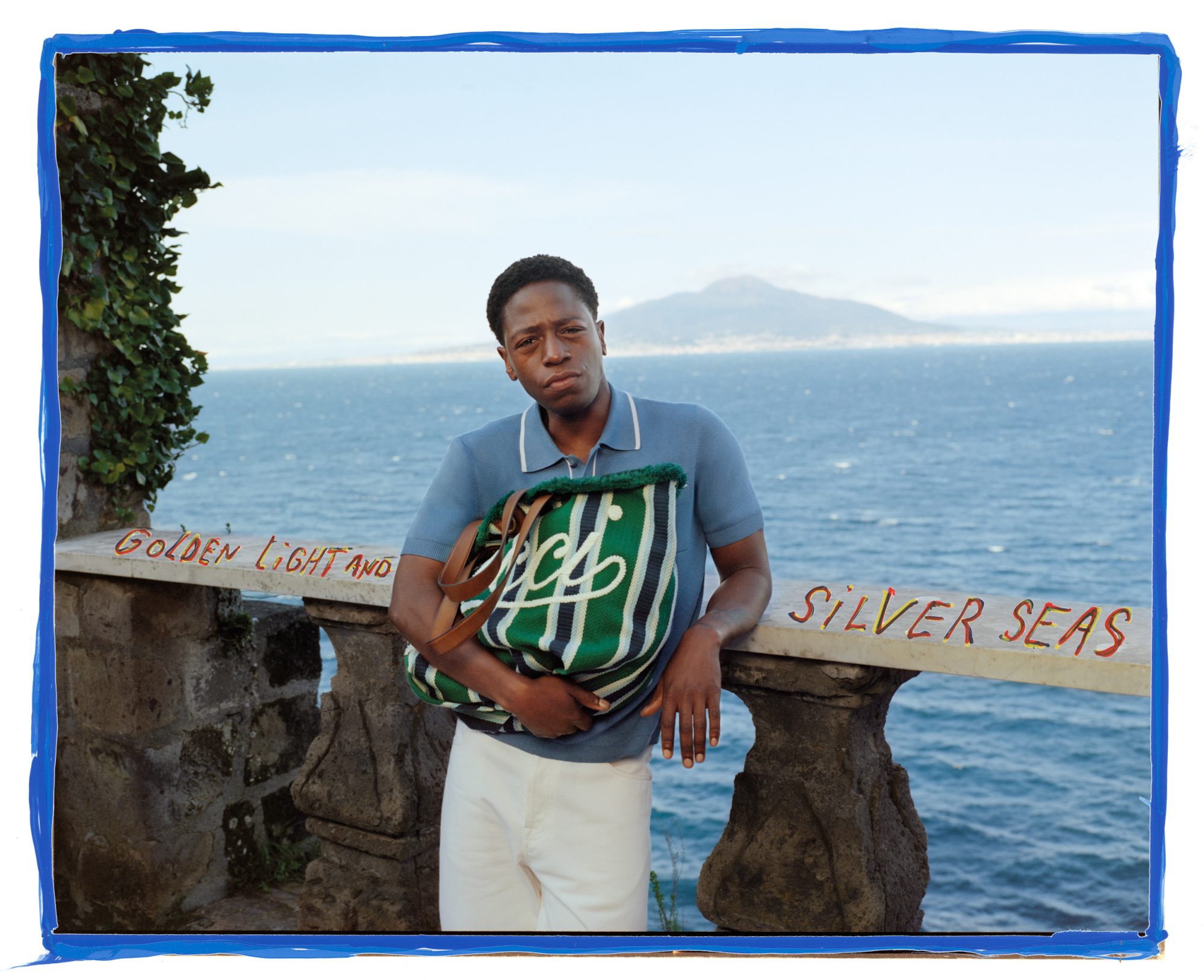 Man poses with a tote bag on a seaside balcony against backdrop of ocean and mountain. He wears a blue polo and white pants.
