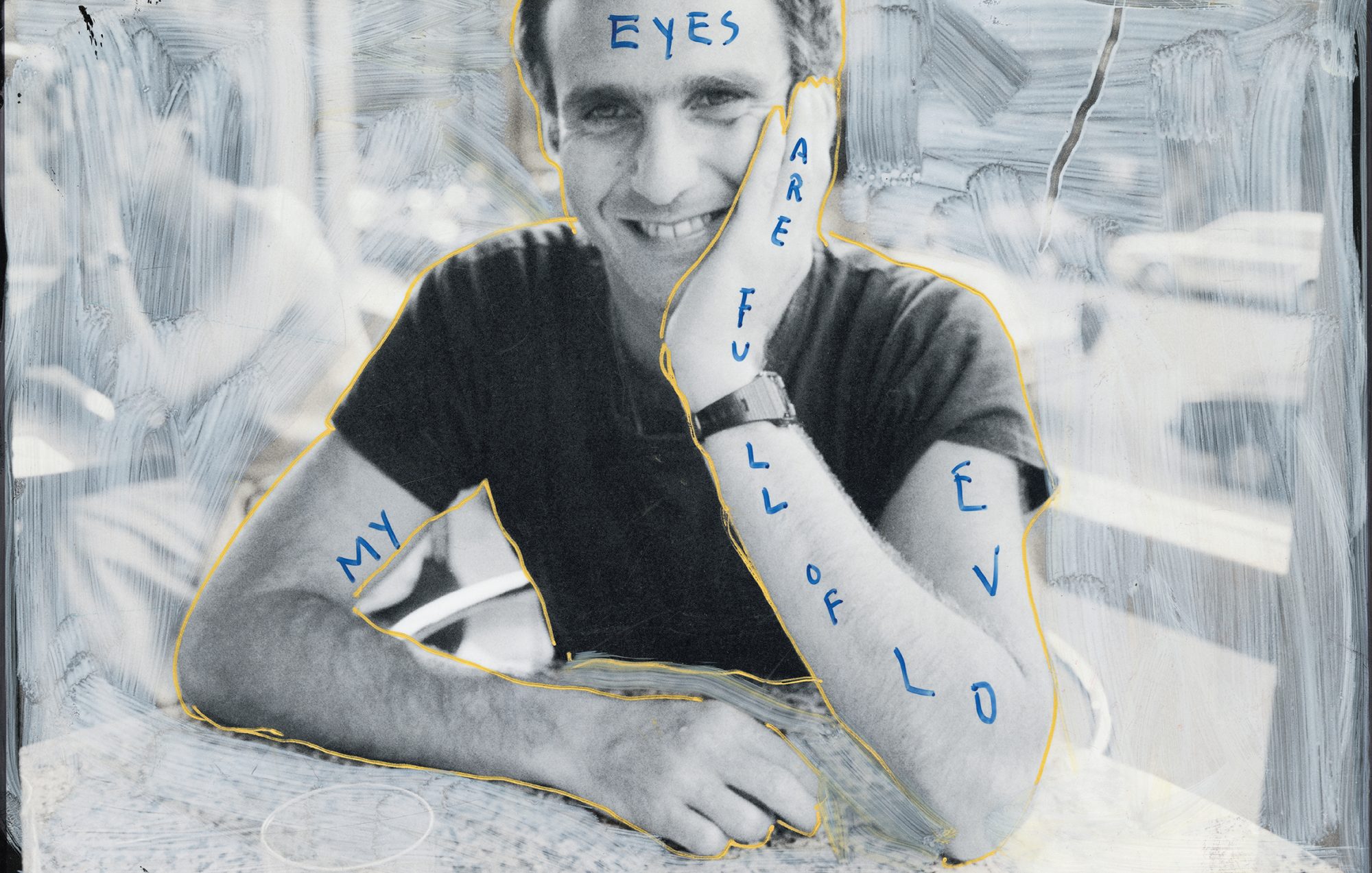 Portrait of a smiling adult male with hand on face and blue ink lettering on his arms and above his head in a black and white image.