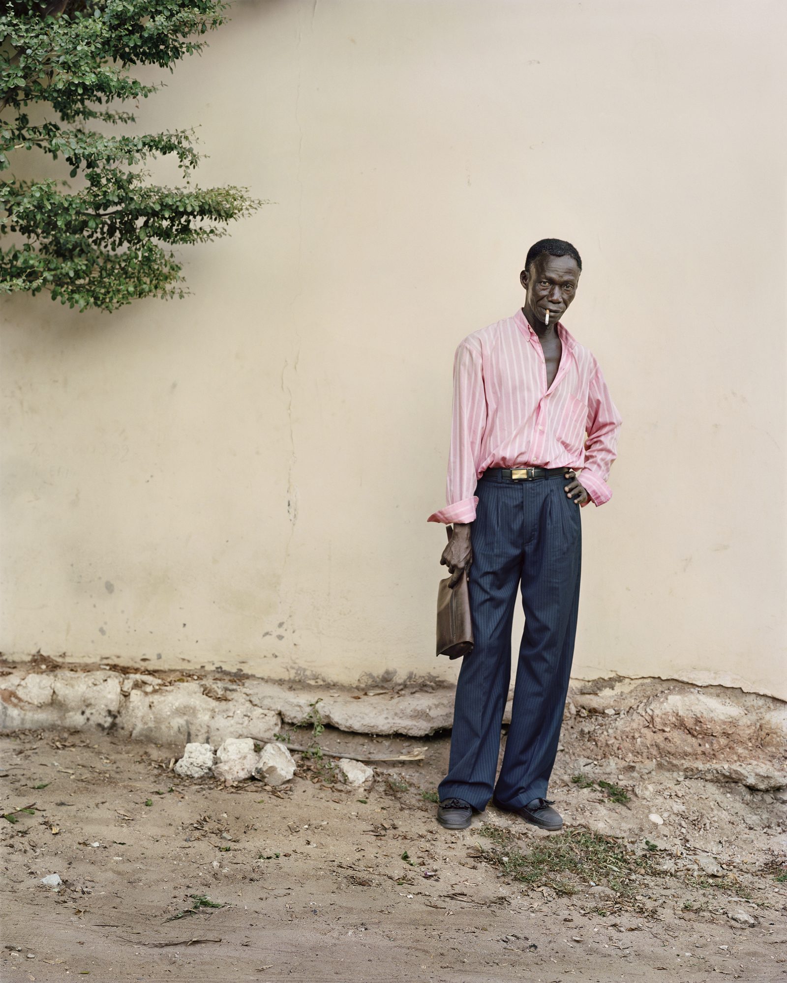 An African man stands confidently outdoors, holding a dark bag, sporting a pink striped shirt and dark pants. He has a cigarette in his mouth.