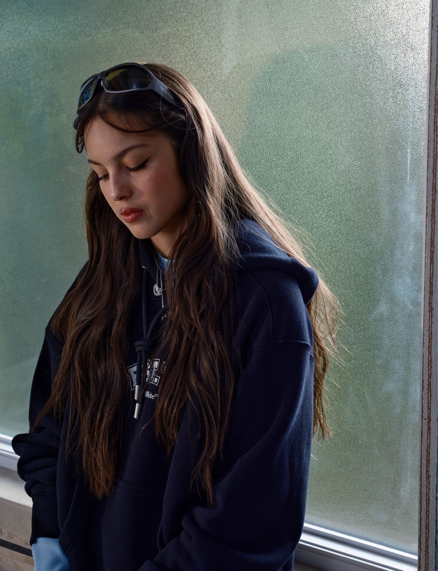 Indoor portrait of a woman with long brown hair wearing a hooded sweatshirt and sunglasses on her head, looking down with a neutral expression.