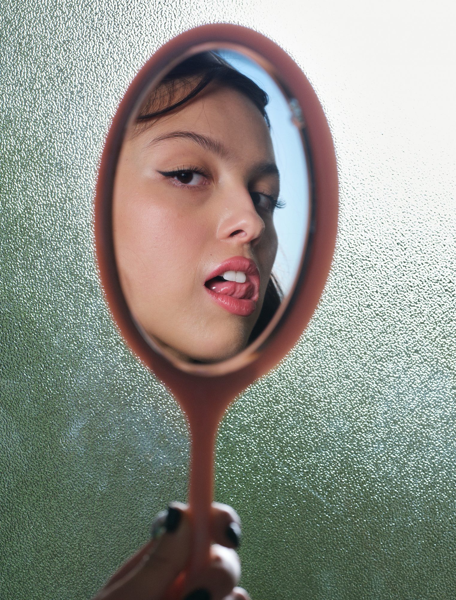 Young woman reflected in a handheld mirror, sticking her tongue out with clear, glossy skin, and wing eyeliner, against textured glass.