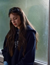 Olivia Rodrigo with long brown hair wearing a dark navy blue hoodie and sunglasses on head, standing near textured glass window