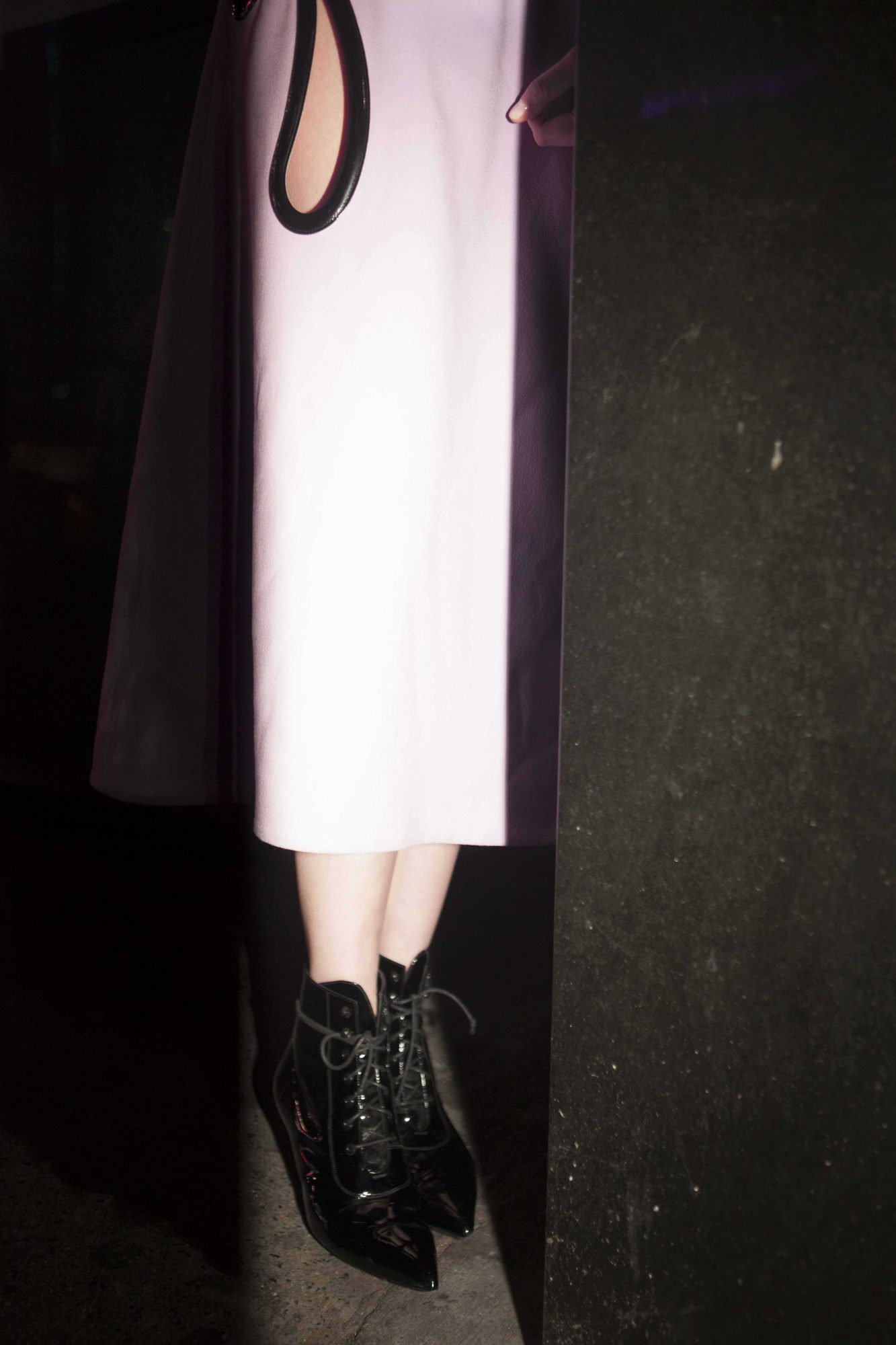 Fashionable woman showcasing black patent leather pointed-toe lace-up boots with a cream-colored dress in a studio setting.