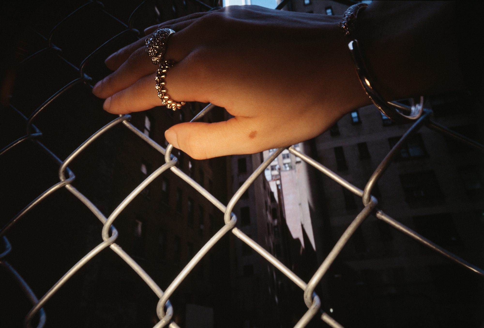 Close-up of hand on chain-link fence with rings and a bracelet, buildings blurred in the background, creating an urban outdoor scene.