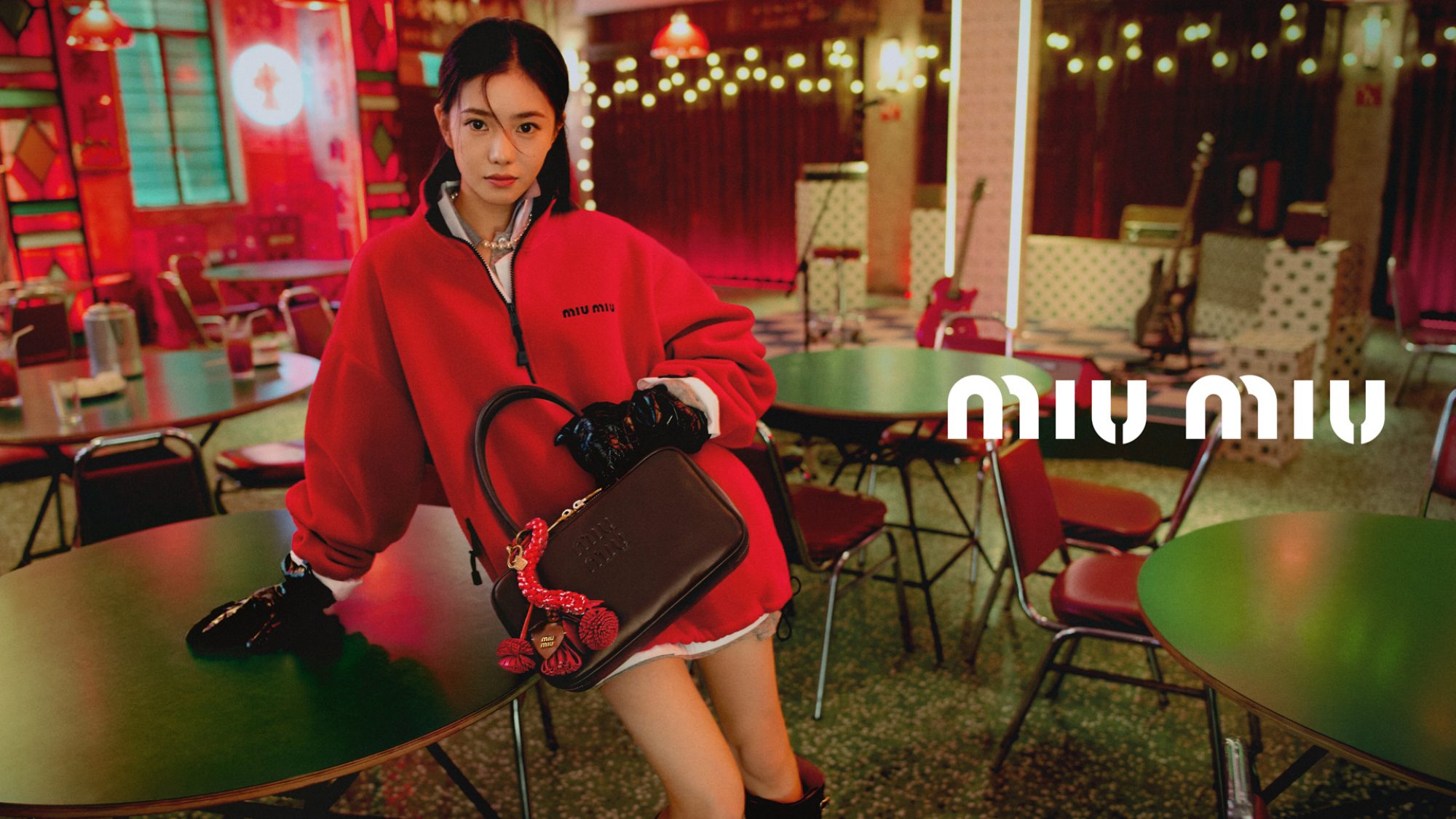 Asian woman models a red Miu Miu sweatshirt and black leather bag in a cafe-style setup with green tables and red chairs.