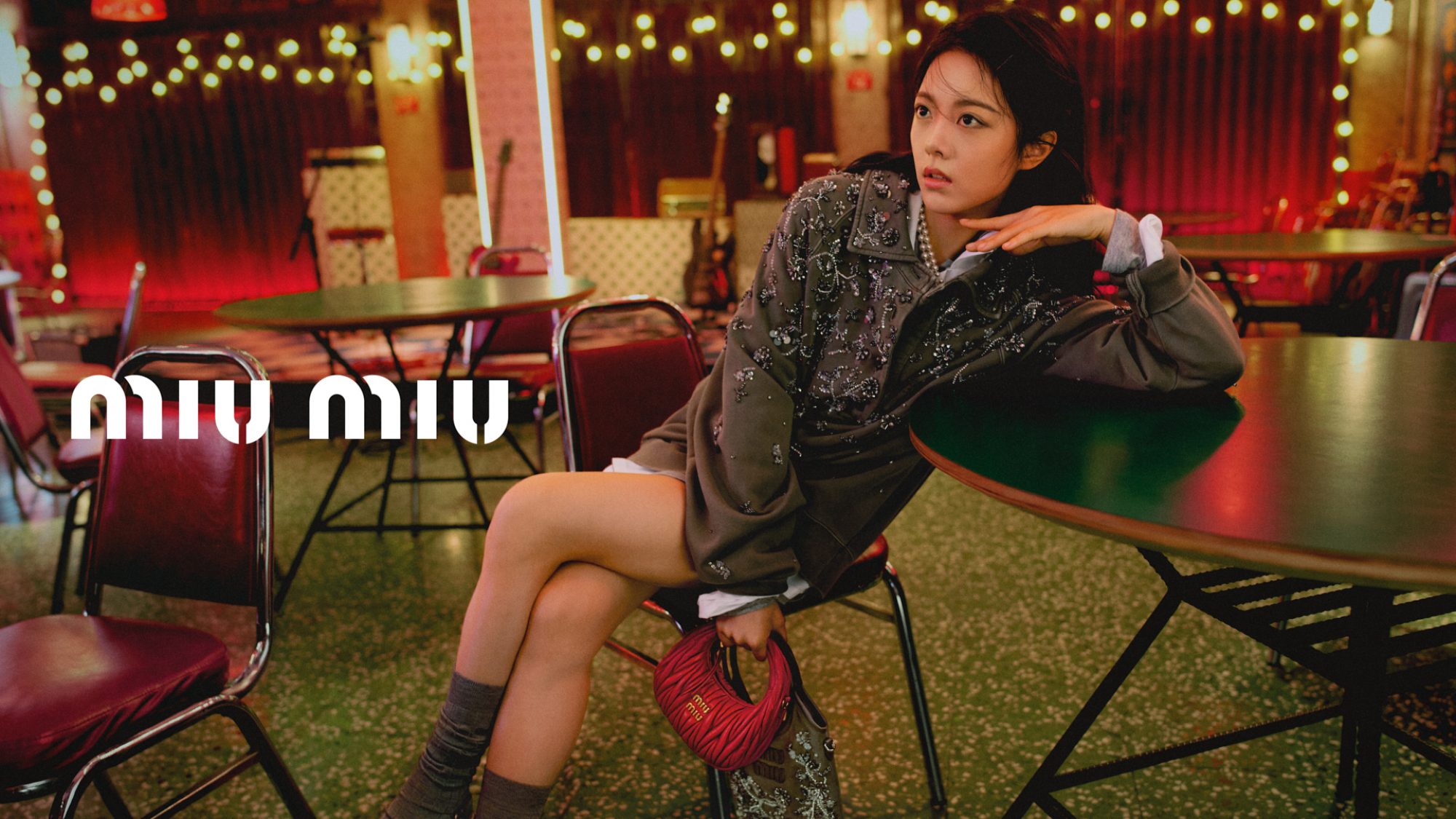 A young woman poses indoors next to a green table in a campaign-style image for Miu Miu, wearing a patterned jacket, and holding a red bag.