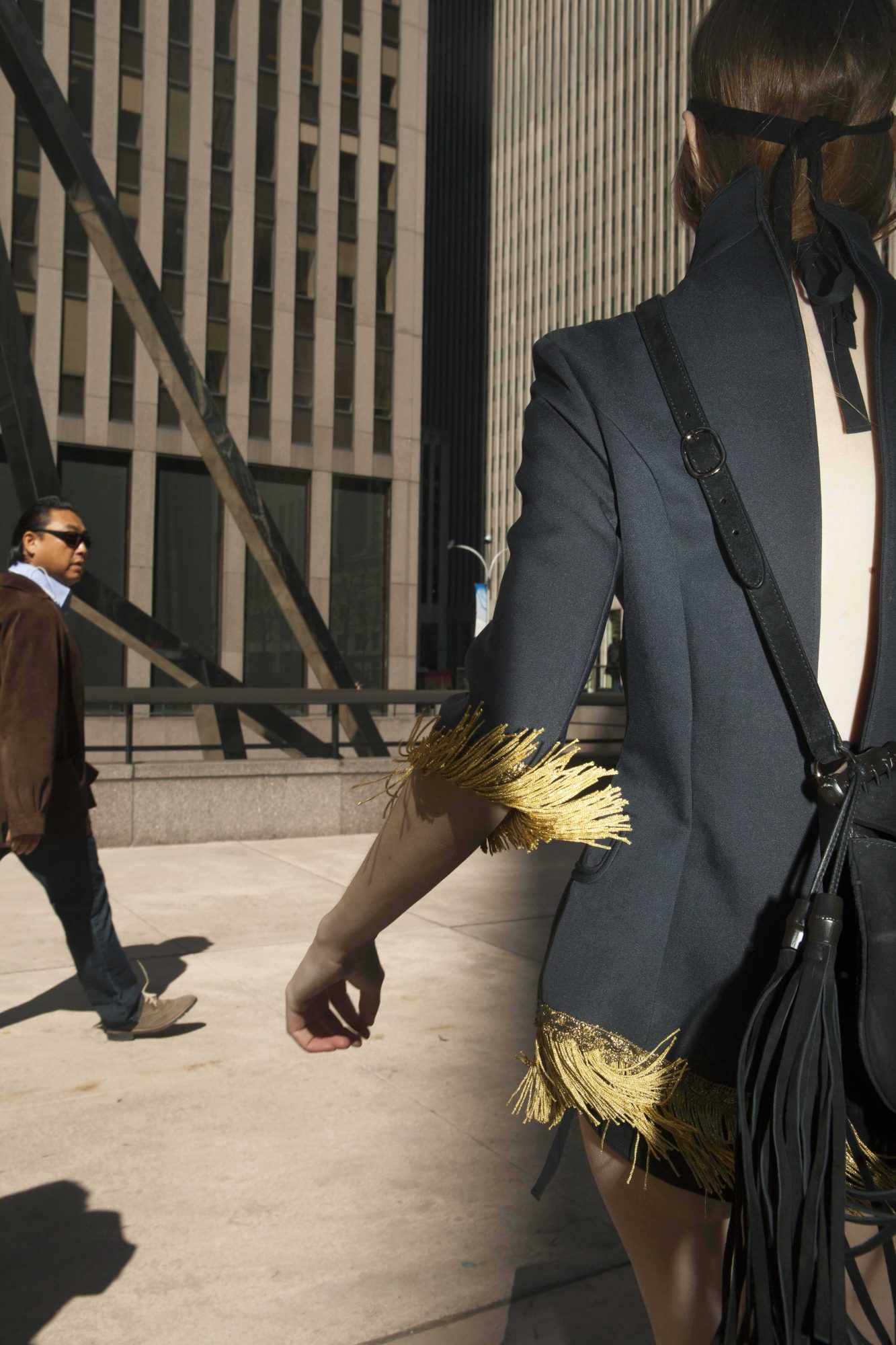 Woman with fringed jacket walks in a city with a man in sunglasses in the background against office buildings and sky.