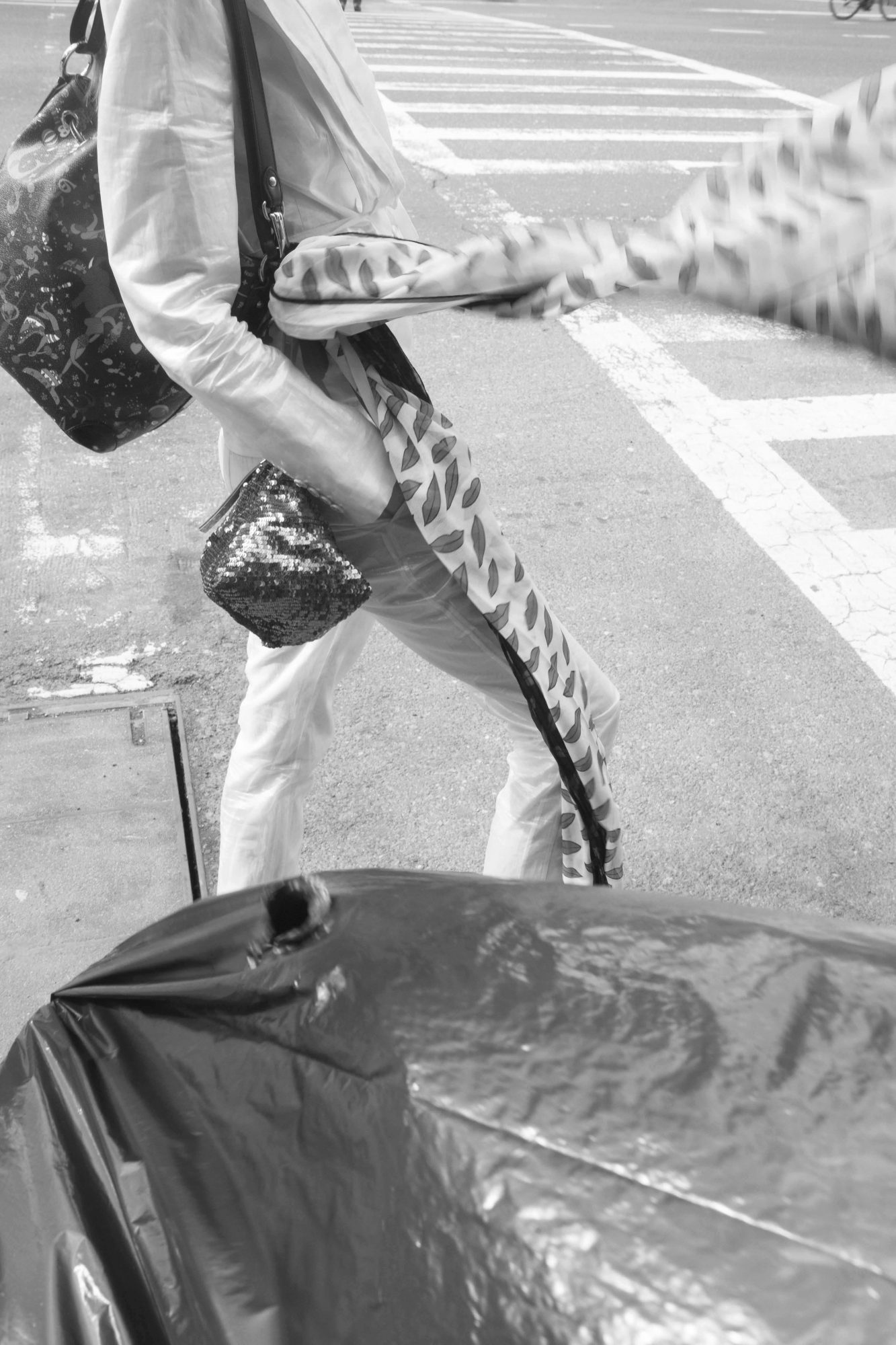 Monochrome street-style image of a person walking with a patterned scarf and bags, captured with a crosswalk and cityscape in the background.