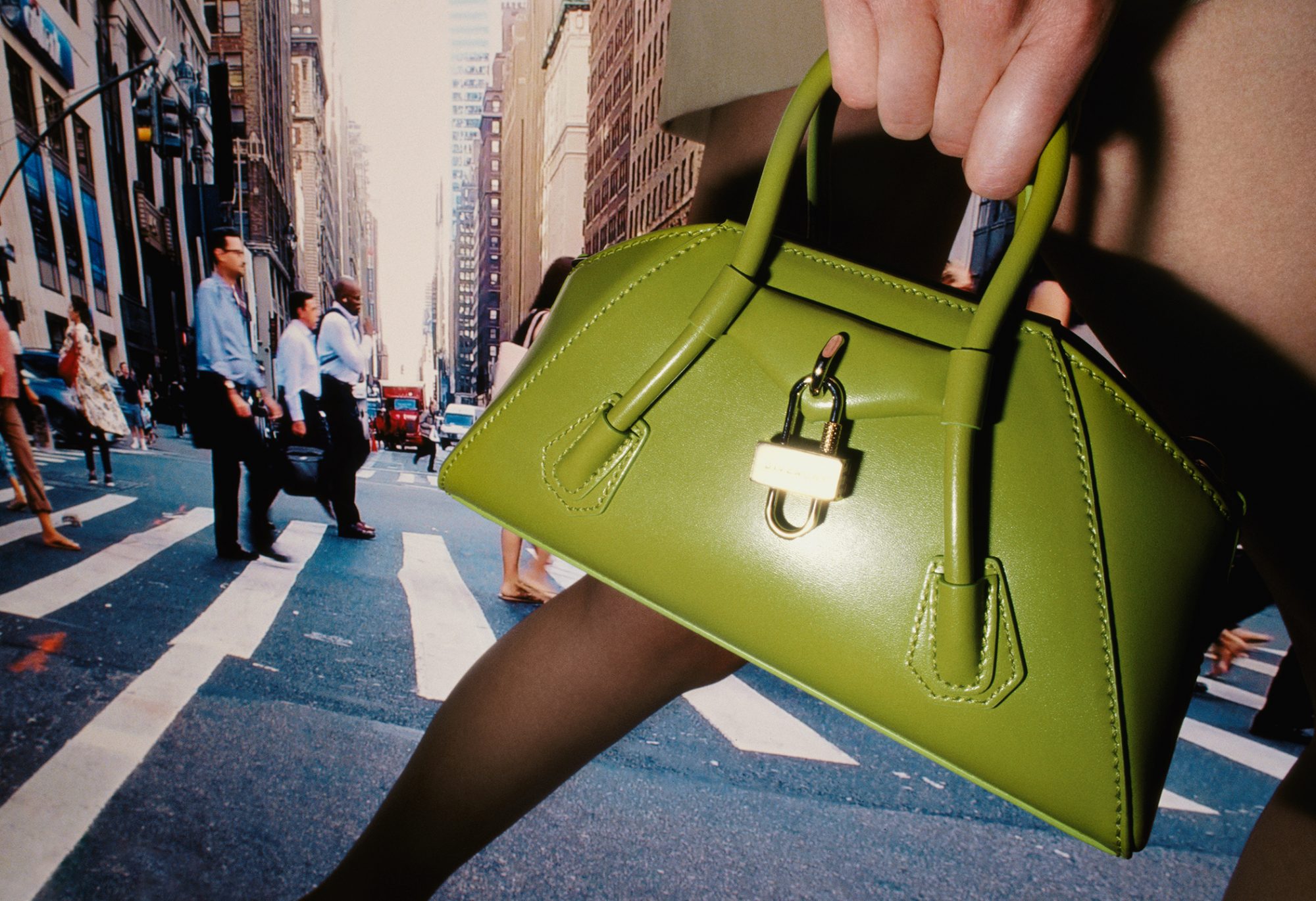 Lime green leather handbag with a padlock in front of a backdrop featuring a busy city street with pedestrians and yellow taxis.
