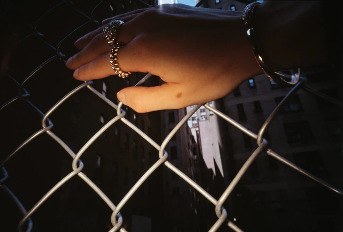 Close-up of a hand with jewelled rings and a bracelet resting against a chain-link fence in New York by photographer Jack Webb