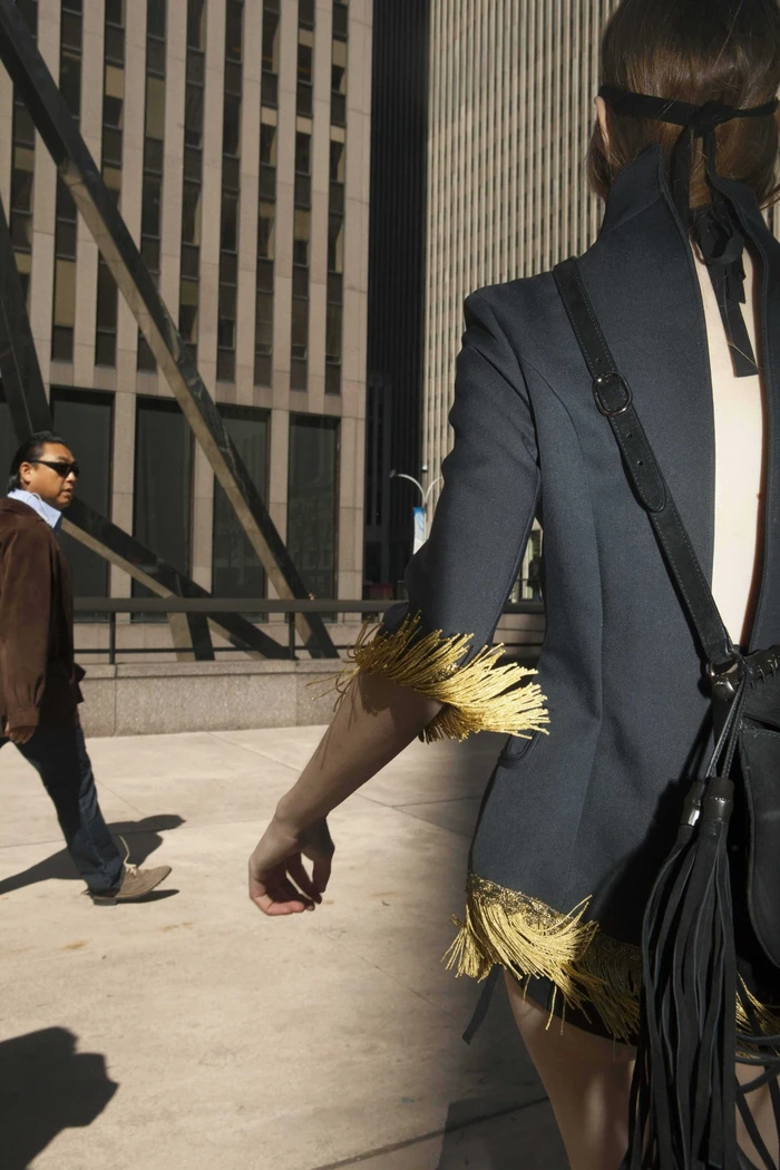 Model in dark jacket with gold fringe and black bag walking on city plaza past tall buildings