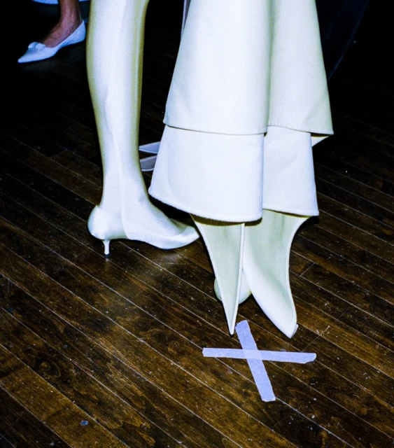 Fashion shoot featuring a woman in a white dress and boots, with attention to the floor marked with a white X.